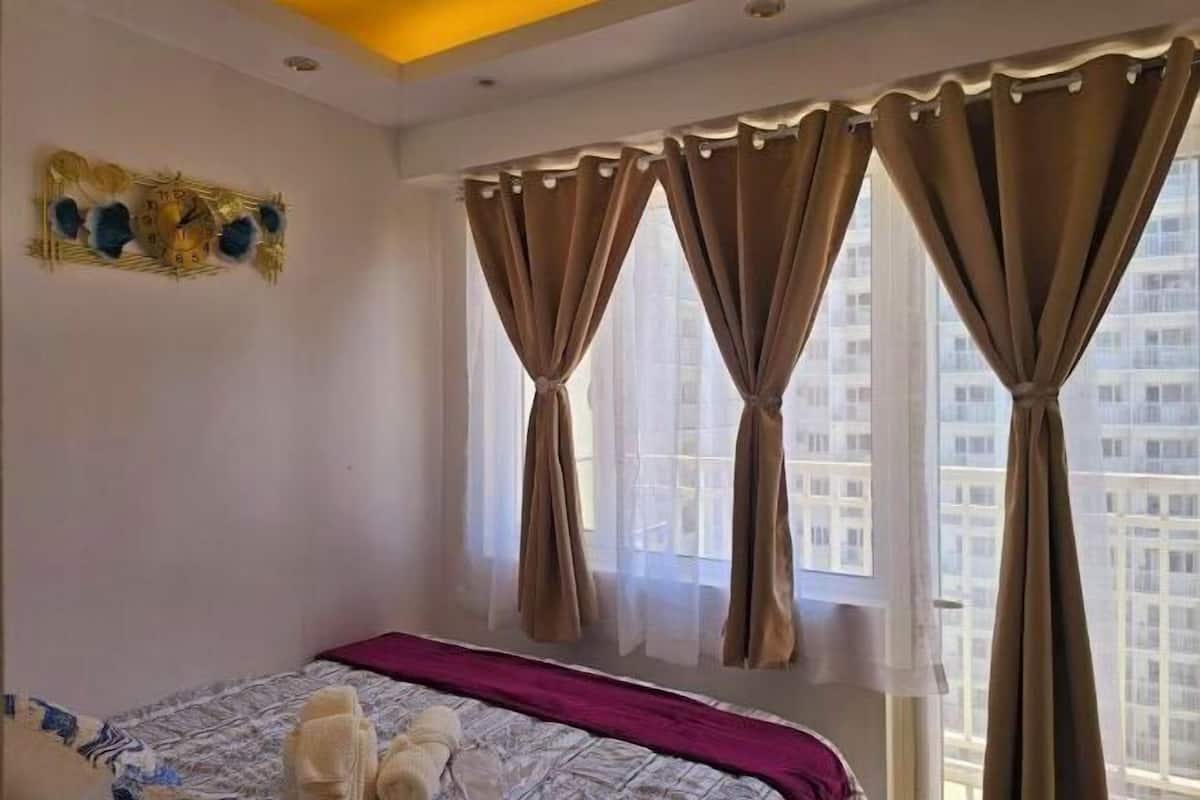 A hotel room with a bed, curtains, a wall clock, and a window with a view of buildings.