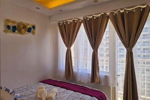 A hotel room with a bed, curtains, a wall clock, and a window with a view of buildings. - Hannah's Crib at Shore Residences with Balcony and Pool View (Pasay)