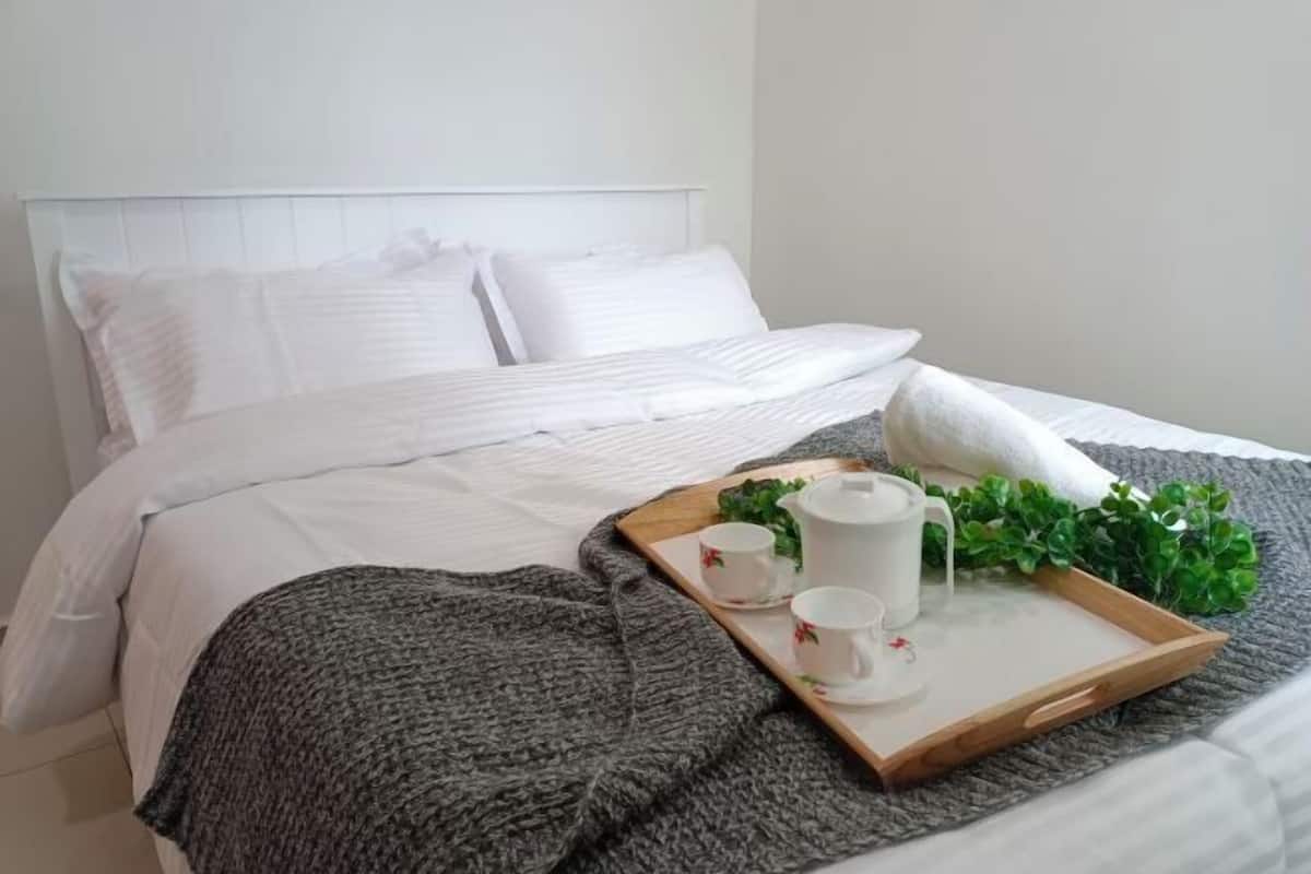 A neatly made bed with white linens, a gray knitted blanket, and a tray with a teapot, cups, and a small plant.