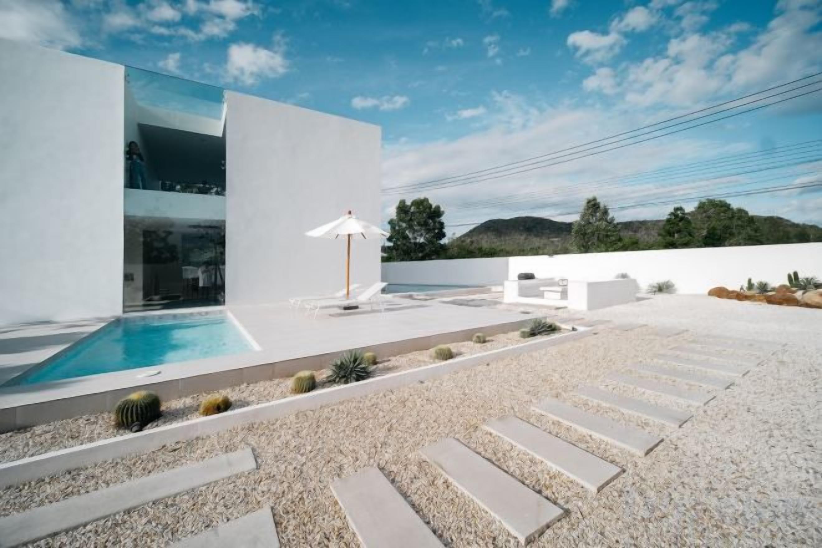 A modern outdoor pool area with a clear sky, a white building, and a seating area with a sun umbrella.