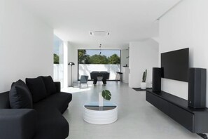 A modern living room with a black sofa, a coffee table with a plant, a flat-screen TV, and a dining area with a table and chairs. - Her Huahin Pool Villa (Hua Hin)