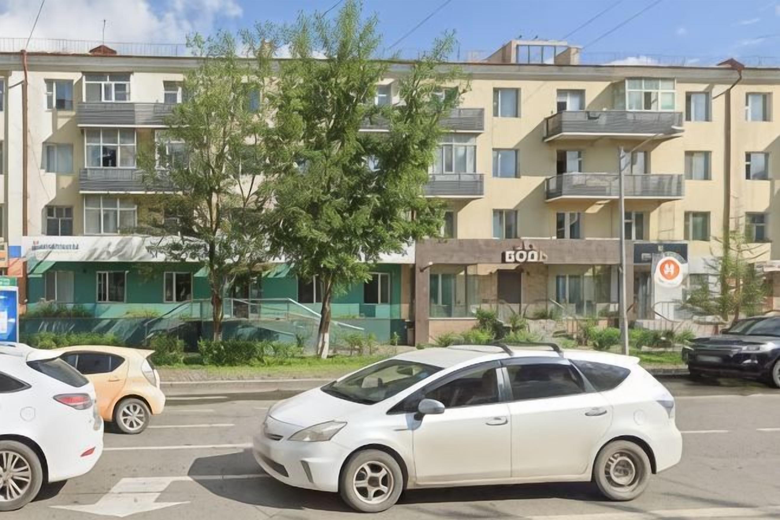 A multi-story building with balconies, a tree in the foreground, and parked cars.