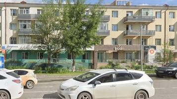 A multi-story building with balconies, a tree in the foreground, and parked cars.
