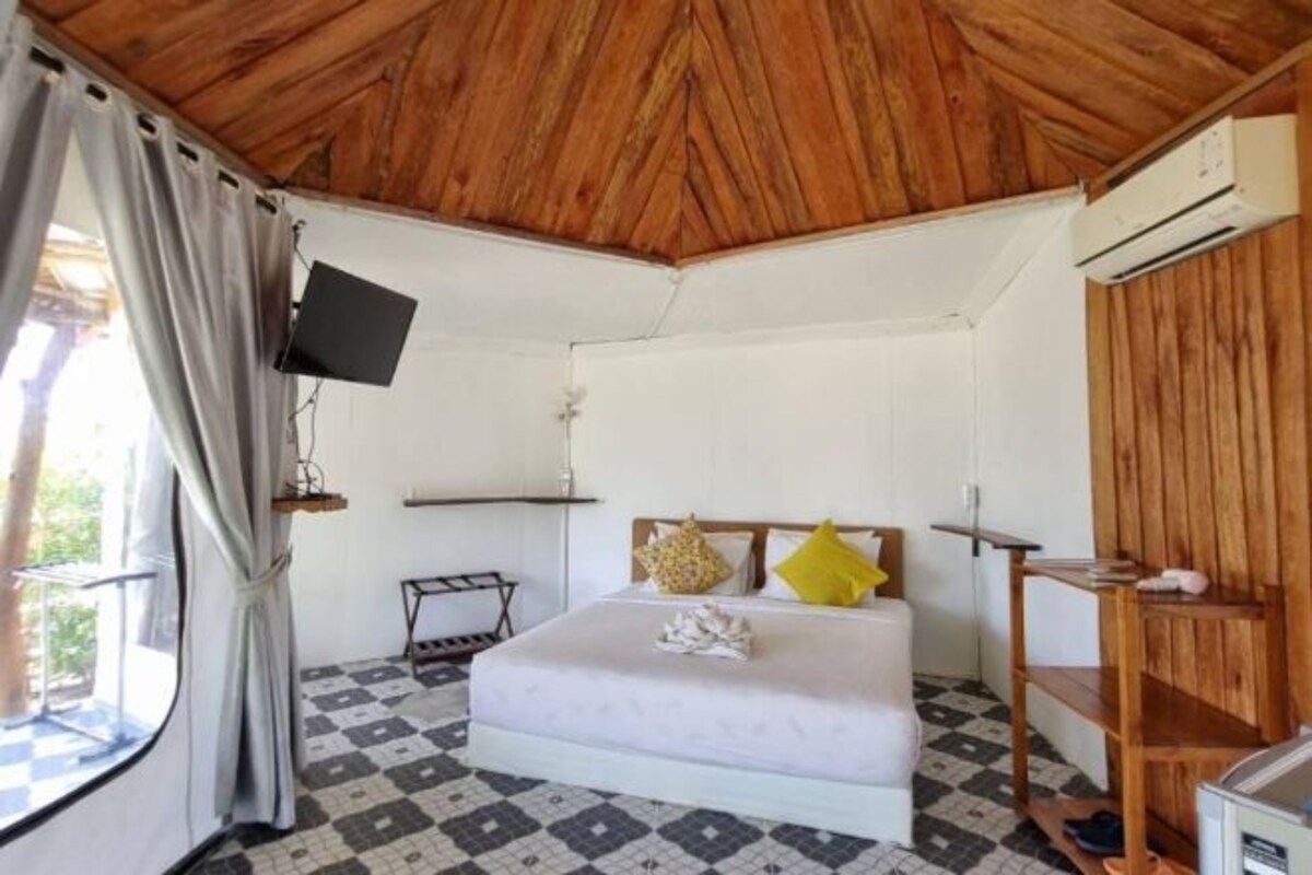 A bedroom with a bed, a mounted TV, a wooden shelf, and a window with curtains.