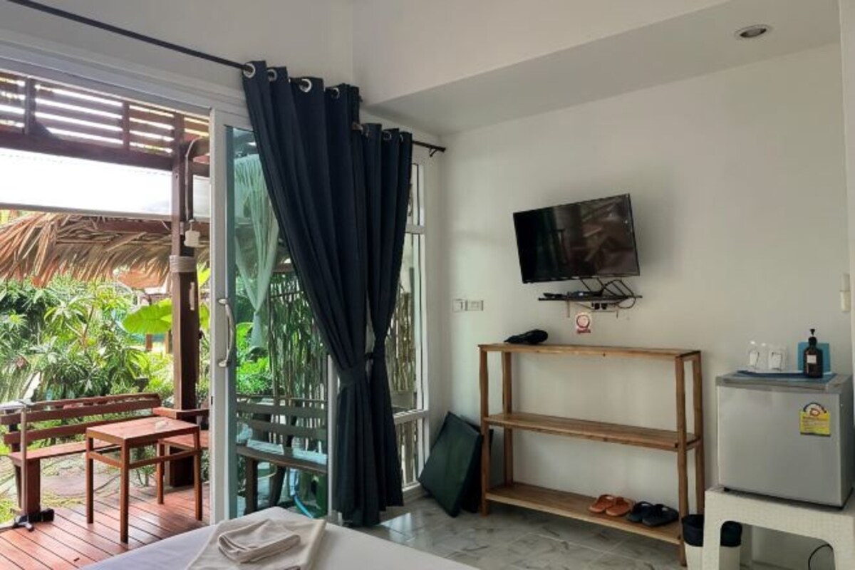 A room with a bed, a TV mounted on the wall, a wooden bench, a small table, and a door leading to an outdoor area with plants.