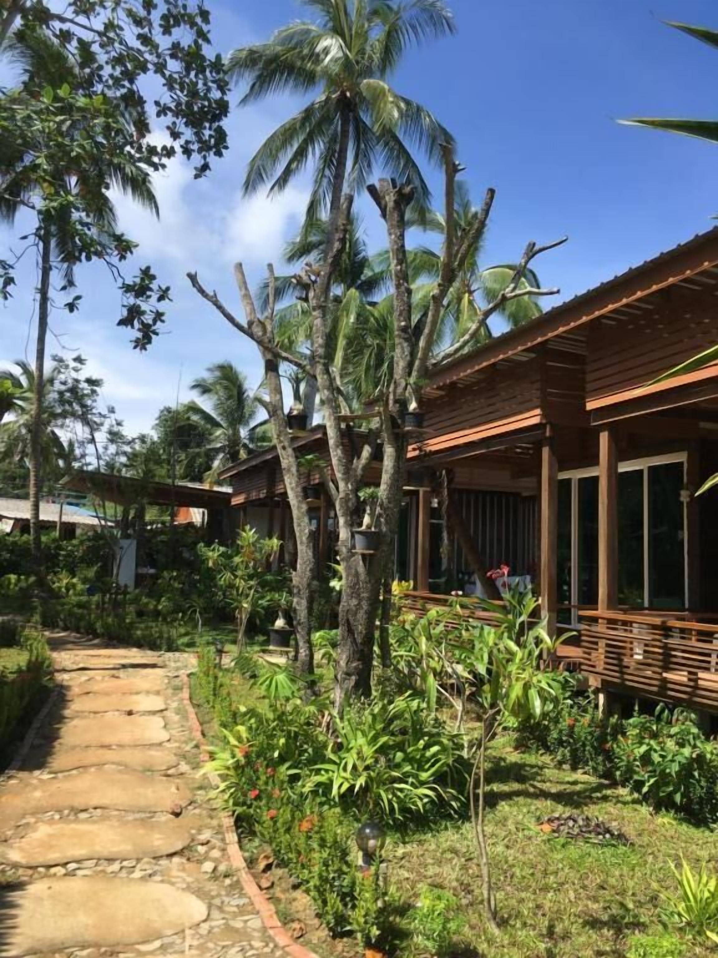 Een tropisch resort met een pad van steen dat leidt naar houten gebouwen, omringd door weelderige begroeiing en palmbomen.