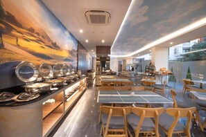 A restaurant with a buffet setup, wooden chairs, and a mural of a sunset on the wall.
