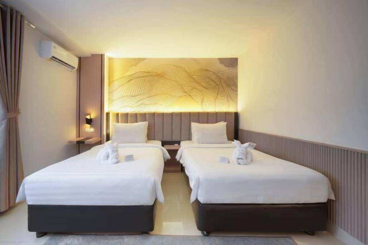 A hotel room with two beds, a wall-mounted air conditioner, a decorative wall panel, and a nightstand with a lamp.