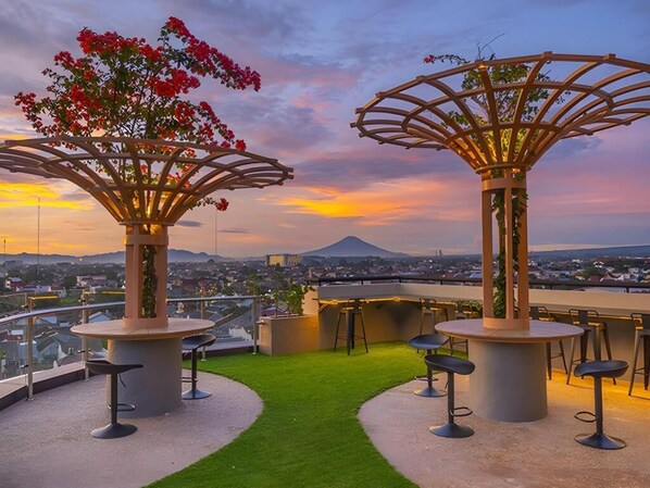 An outdoor seating area with round tables and chairs, surrounded by green artificial grass, and overlooking a cityscape with a mountain in the distance.