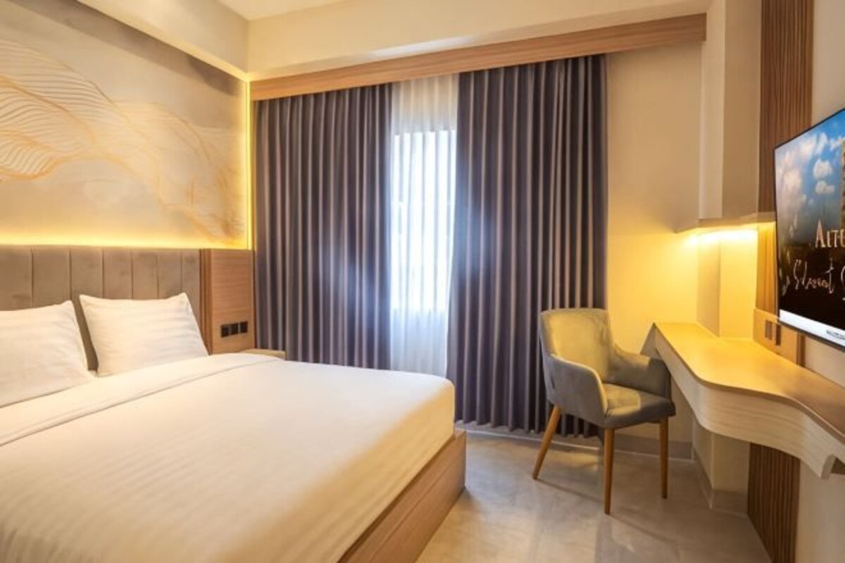 A hotel room with a bed, a desk with a chair, a TV, and a large window with curtains.