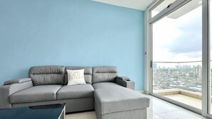 A grey sectional sofa with a white pillow, a small table, and a large window with a city view. - KSL D'Esplanade 2Bedrooms 7-8pax Shopping Heaven (Johor Bahru)