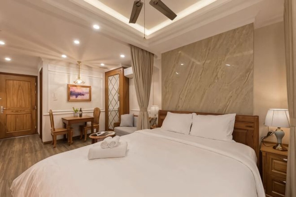 A hotel room with a large bed, a desk with a chair, a small table, a ceiling fan, and a large mirror.