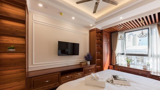 A hotel room with a large bed, a flat-screen TV mounted on the wall, wooden paneling on the walls, a window with curtains, and a ceiling fan. - Woody House Boutique (Ho Chi Minh Municipality)