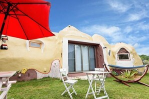 Ein außergewöhnliches Haus mit geschwungener Fassade, einer Schiebetür aus Glas, einer Hängematte und einem roten Sonnenschirm.