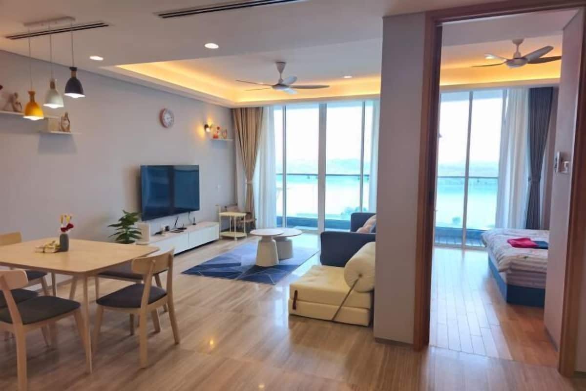 A modern hotel room with a dining area, a sofa, a TV, and a balcony with a view of the sea.