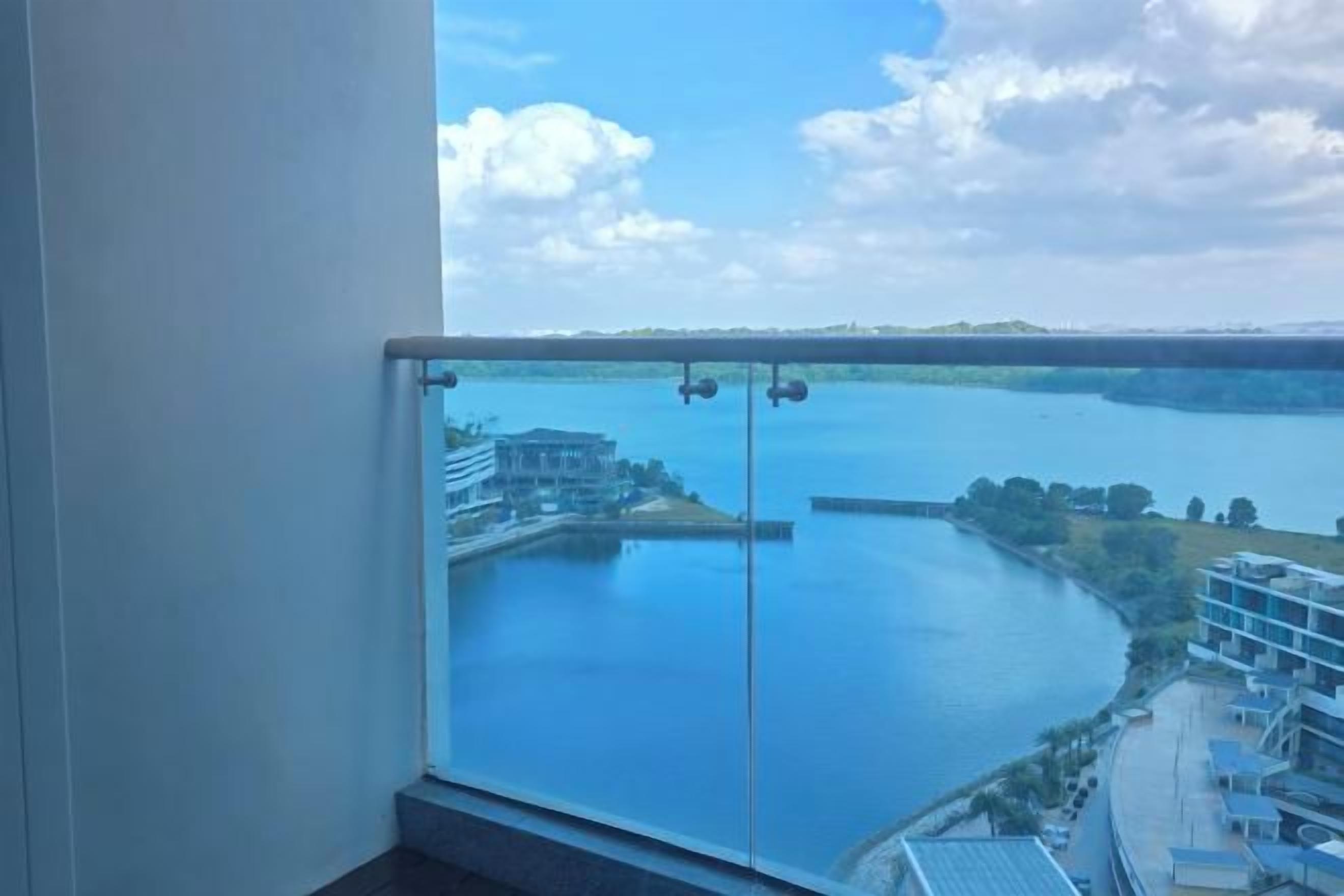 A balcony with a glass railing overlooking a body of water, buildings, and a clear sky.