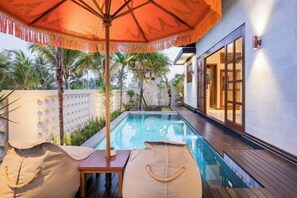 Un espace piscine avec des chaises longues et un parasol, entouré de palmiers et d’un mur blanc.
