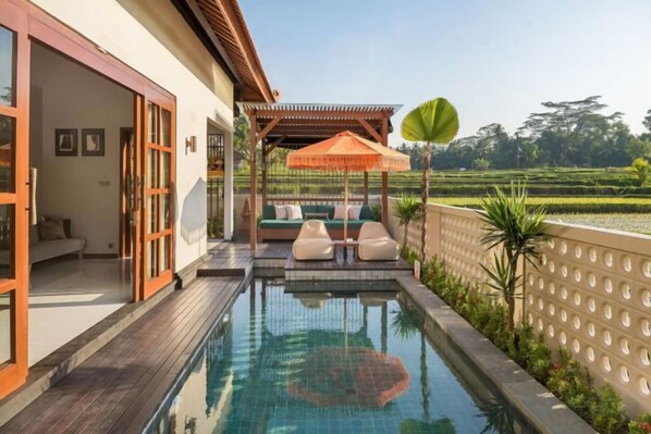 A swimming pool with a wooden deck, a covered seating area with cushions, and a view of a landscaped garden. - Dijero at Umah Bali (Bali)