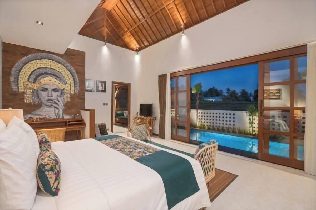 A bedroom with a large bed, a desk, a chair, a view of a pool, and a mural on the wall.