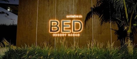 A sign for "BED RESORT SADAO" with illuminated letters against a wooden background, surrounded by grass and palm trees.