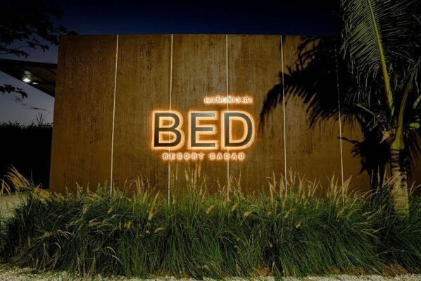 A sign for "BED RESORT SADAO" with illuminated letters against a wooden background, surrounded by grass and palm trees. - Bed Resort Sadao (Sadao)