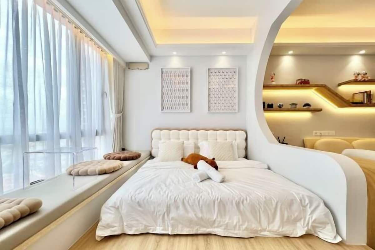 A modern bedroom with a curved headboard, a large window with sheer curtains, a bedside table, and a built-in shelf with decorative items.