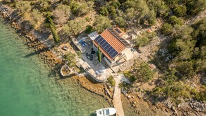 An aerial view of a coastal property with a house featuring a solar panel roof, a dock with a boat, and a clear view of the surrounding rocky shoreline and vegetation. - ZadarVillas Off-Grid Private Island Retreat (Sali)