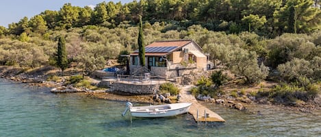 A house with a stone facade, a red-tiled roof, and solar panels. A small boat is docked in the water in front of the house.