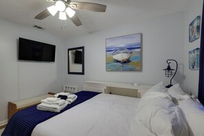 A hotel room with a bed, a ceiling fan, a painting of a boat, and a wall-mounted TV.