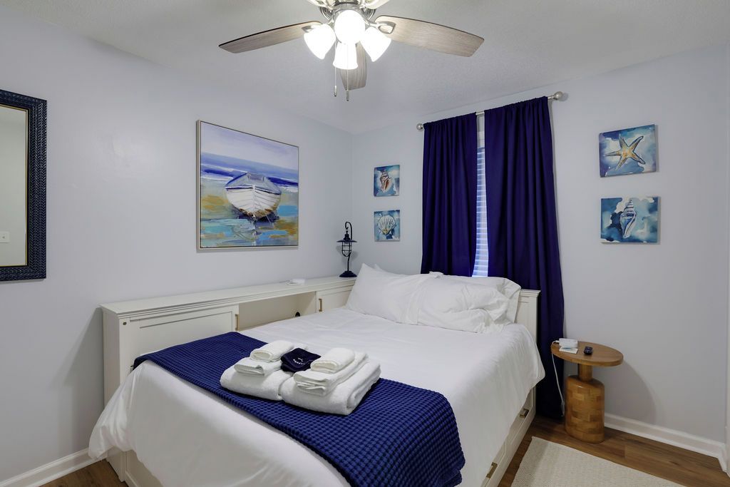 A hotel room with a bed, a ceiling fan, a nightstand, a small table, and artwork on the walls.