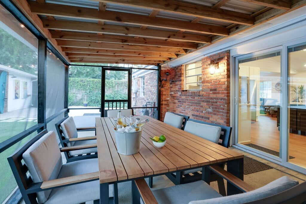 Een terras met een houten tafel en stoelen, een bakstenen muur en grote ramen.