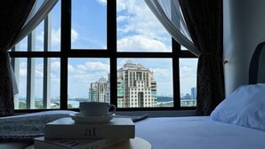 A hotel room with a large window offering a view of a cityscape, a bed, a bedside table with a cup and a book, and a pillow. - Putrajaya Antara Residence Air-Con Suite Facing Lake View (Putrajaya)