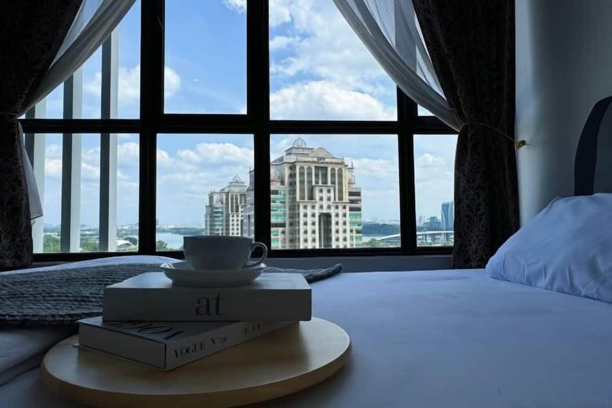 A hotel room with a large window offering a view of a cityscape, a bed, a bedside table with a cup and a book, and a pillow.