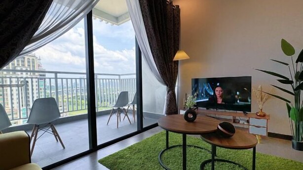 A modern living room with a large TV, a round wooden table, a green rug, and a balcony with a view of buildings. - Putrajaya Antara Residence Air-Con Suite Facing Lake View (Putrajaya)
