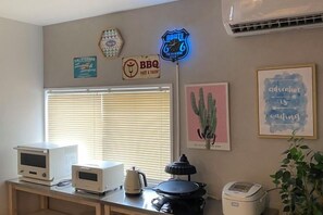 A kitchen with a microwave, kettle, and a coffee maker on the counter. There are wall decorations including a neon sign, a cactus painting, and a framed quote. - HOTEL YANBARUNIA (Nago)