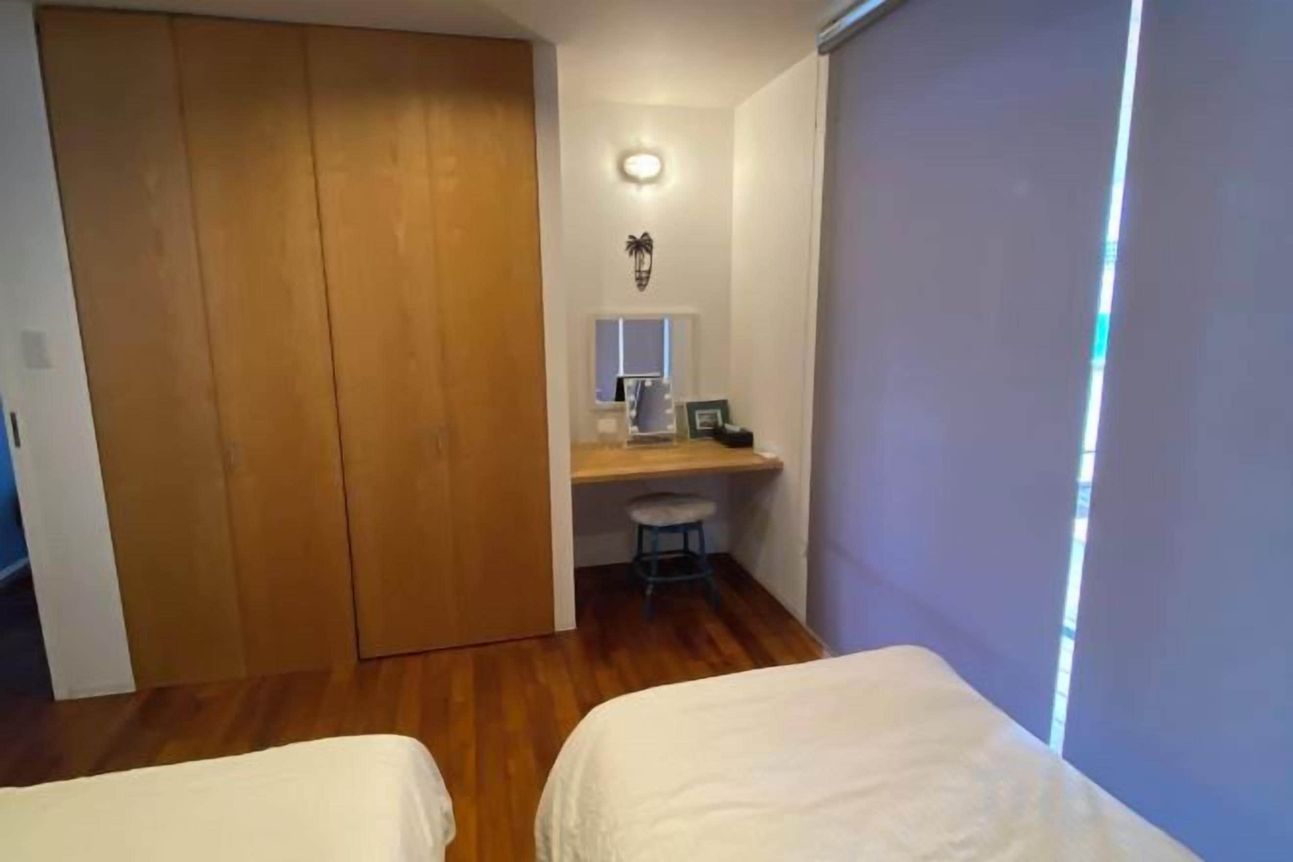 A bedroom with a wooden wardrobe, a desk with a chair, and two beds.