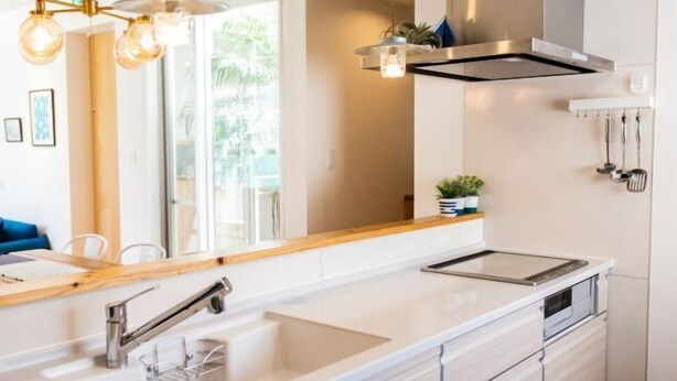 Cozinha moderna com bancada branca, pia de aço inoxidável e coifa. Há uma janela que oferece luz natural e vista para o verde lá fora.