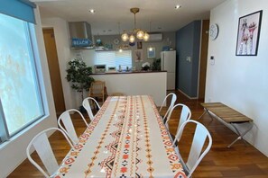 A dining area with a long table, white chairs, and a patterned tablecloth. There is a kitchen in the background with a sign that says "BEACH". - HOTEL YANBARUNIA (Nago)