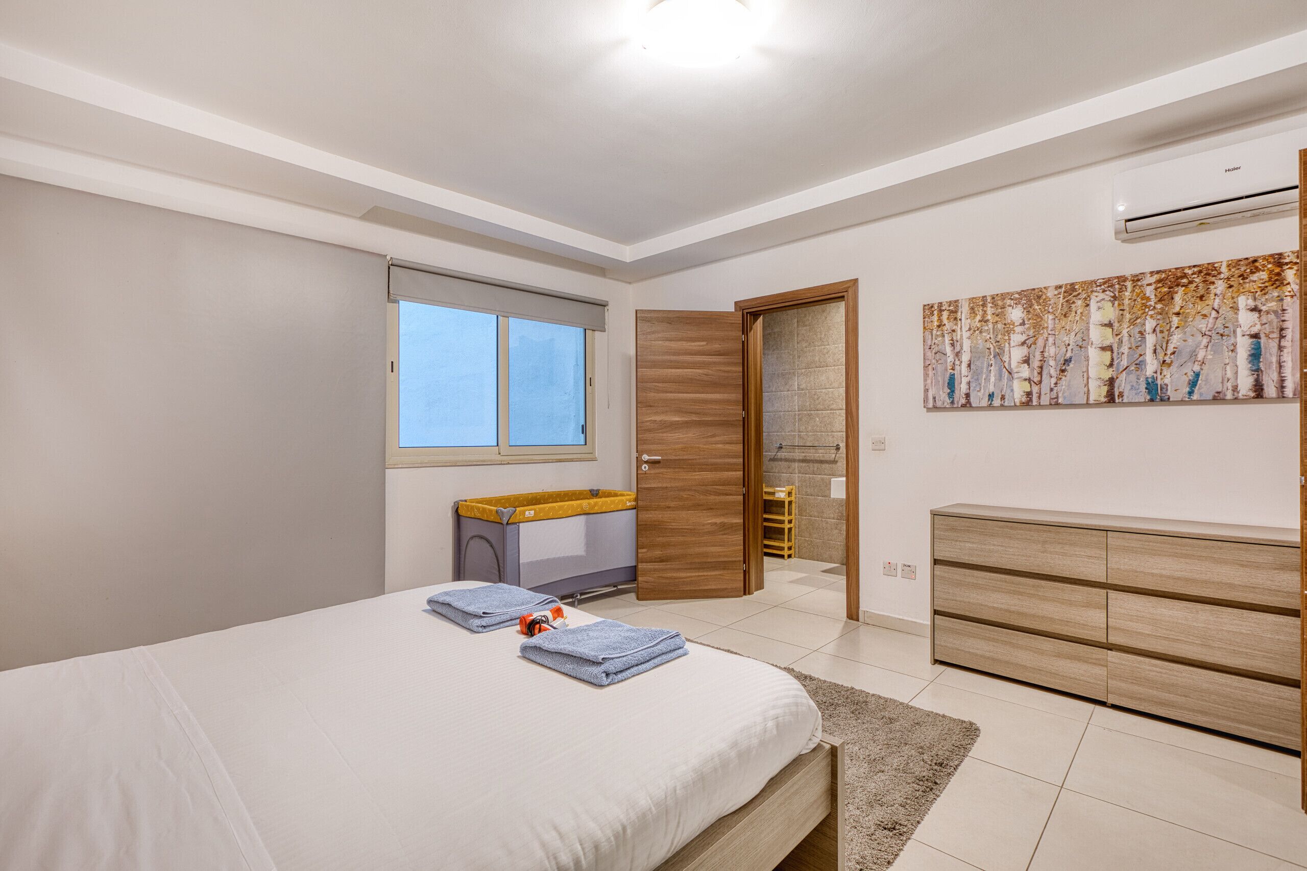 A bedroom with a bed, a wooden dresser, a window, and a painting on the wall.
