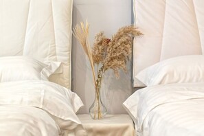 Una cama con sábanas blancas, un florero con plantas secas y una pared de color claro.