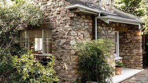 Une maison en pierre avec une cheminée, un petit porche et un jardin où poussent différentes espèces de plantes.