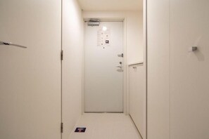 A narrow hallway with a white door at the end, a small sign on the floor, and a light switch on the wall. - Leclerc Higashi-Shinjuku (Shinjuku)