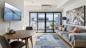 A modern living room with a flat-screen TV, a sofa, a dining table, and a large window. - Comfortable 2BR Retreat With Balcony & Parking (Melbourne)
