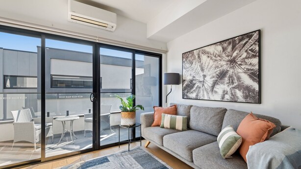 A modern living room with a grey sofa, a large abstract painting, and a sliding glass door leading to an outdoor seating area. - Comfortable 2BR Retreat With Balcony & Parking (Melbourne)