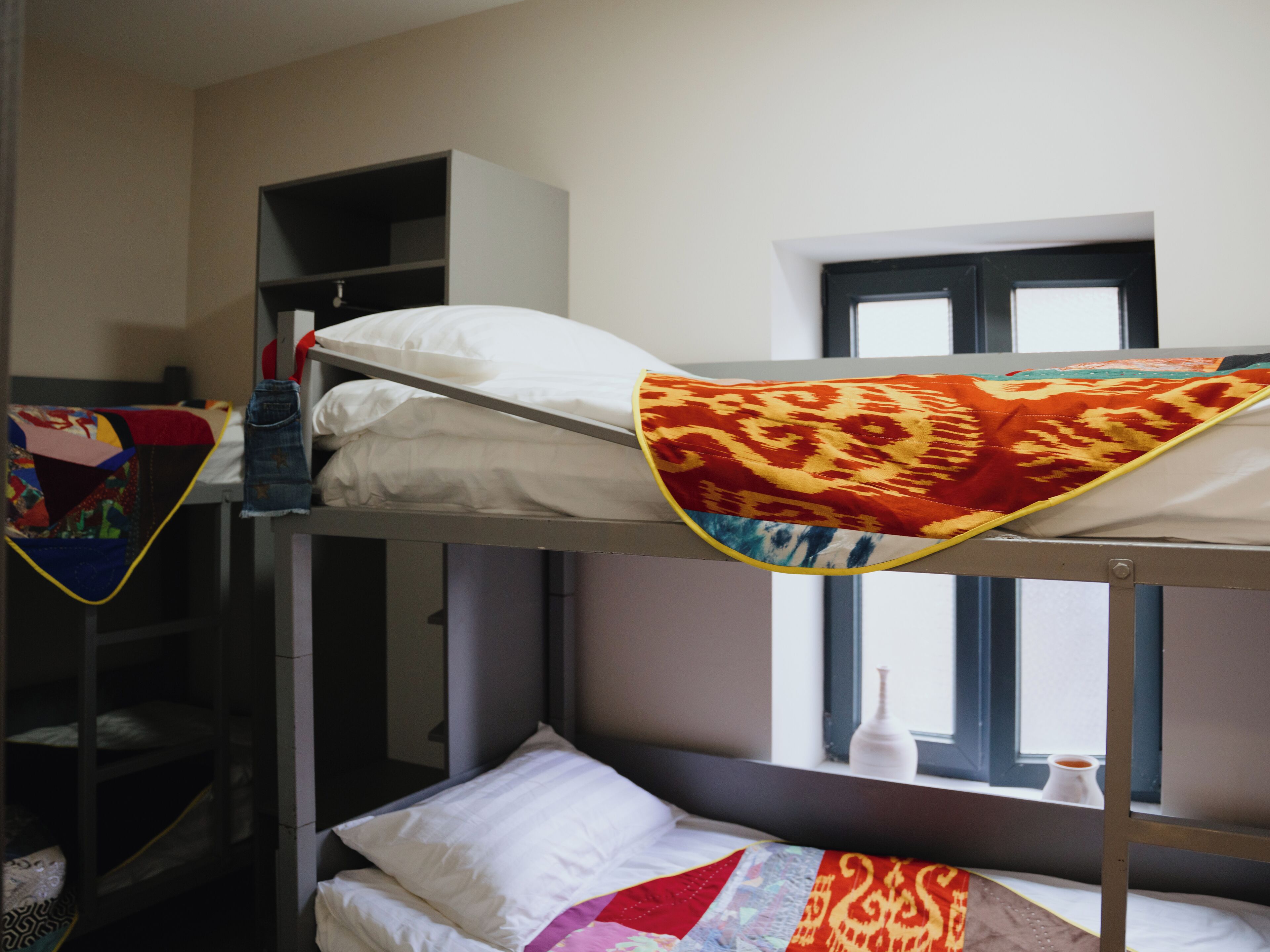 A bunk bed room with a window, a shelf with a red and yellow patterned blanket, and a white pillow.