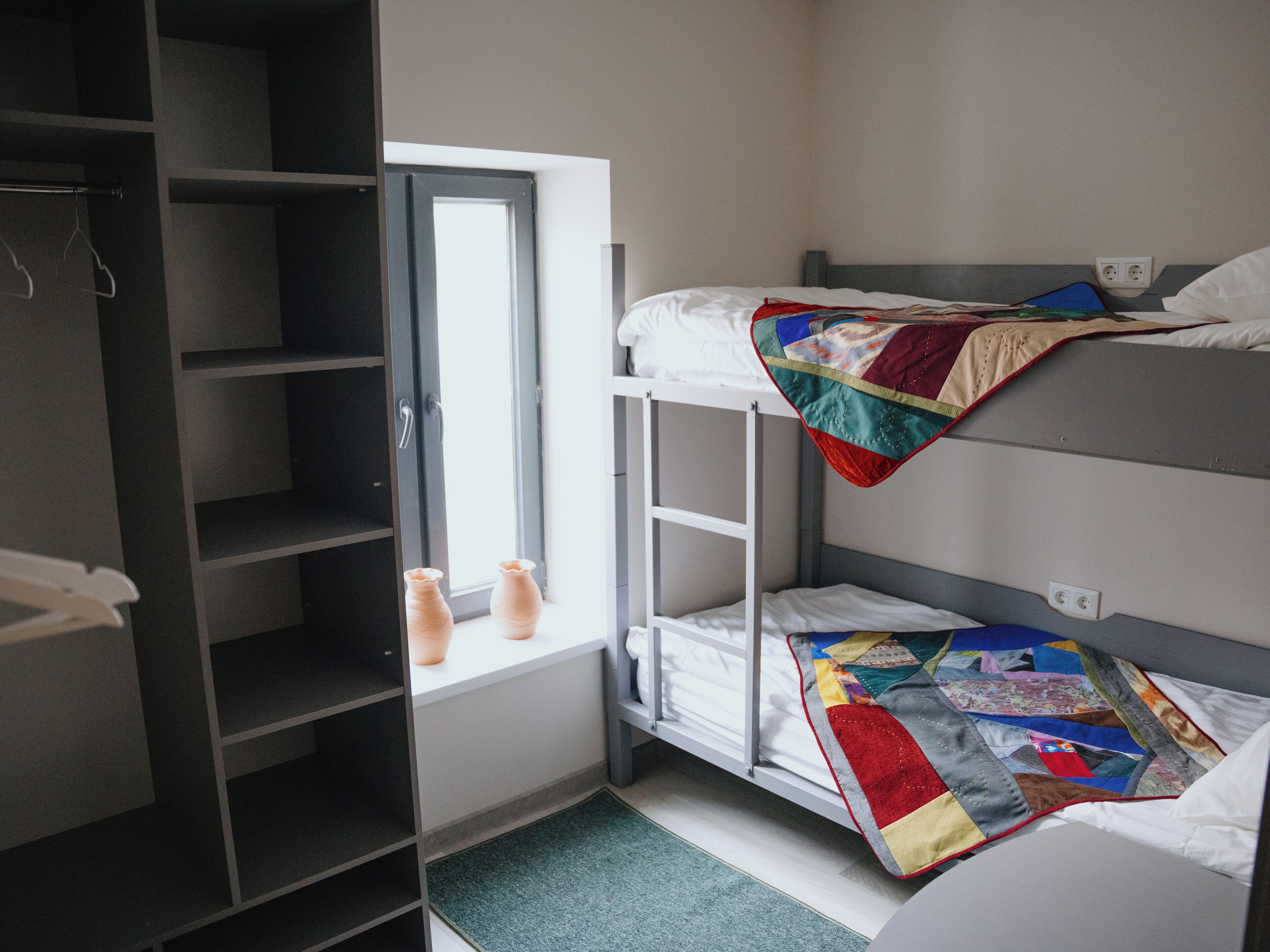 A bunk bed room with a window, a shelf with two vases, and a colorful quilt.