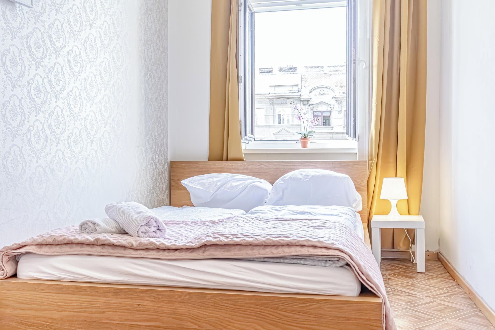 Un lit bien fait, avec une couverture rose et des oreillers blancs, une petite table de chevet avec une lampe et une fenêtre avec des rideaux.