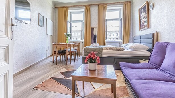 Ein gemütliches Hotelzimmer mit einem Bett, einem violetten Sofa, einem Esstisch mit Stühlen und einem kleinen Tisch mit Blumen.