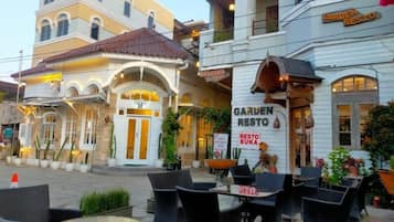An outdoor seating area with wicker furniture and a table, surrounded by buildings with signs for 'GARDEN RESTO' and 'BUKA RESTO'.
