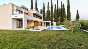 Una casa moderna de dos pisos con piscina, césped cuidado y árboles altos al fondo.